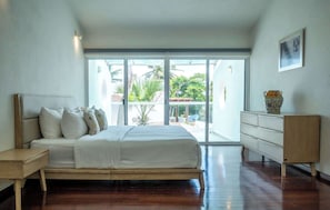 Ein Schlafzimmer mit Bett, Schminktisch, Nachttisch und einer großen Glasschiebetür, die auf einen Balkon mit viel Grün führt.