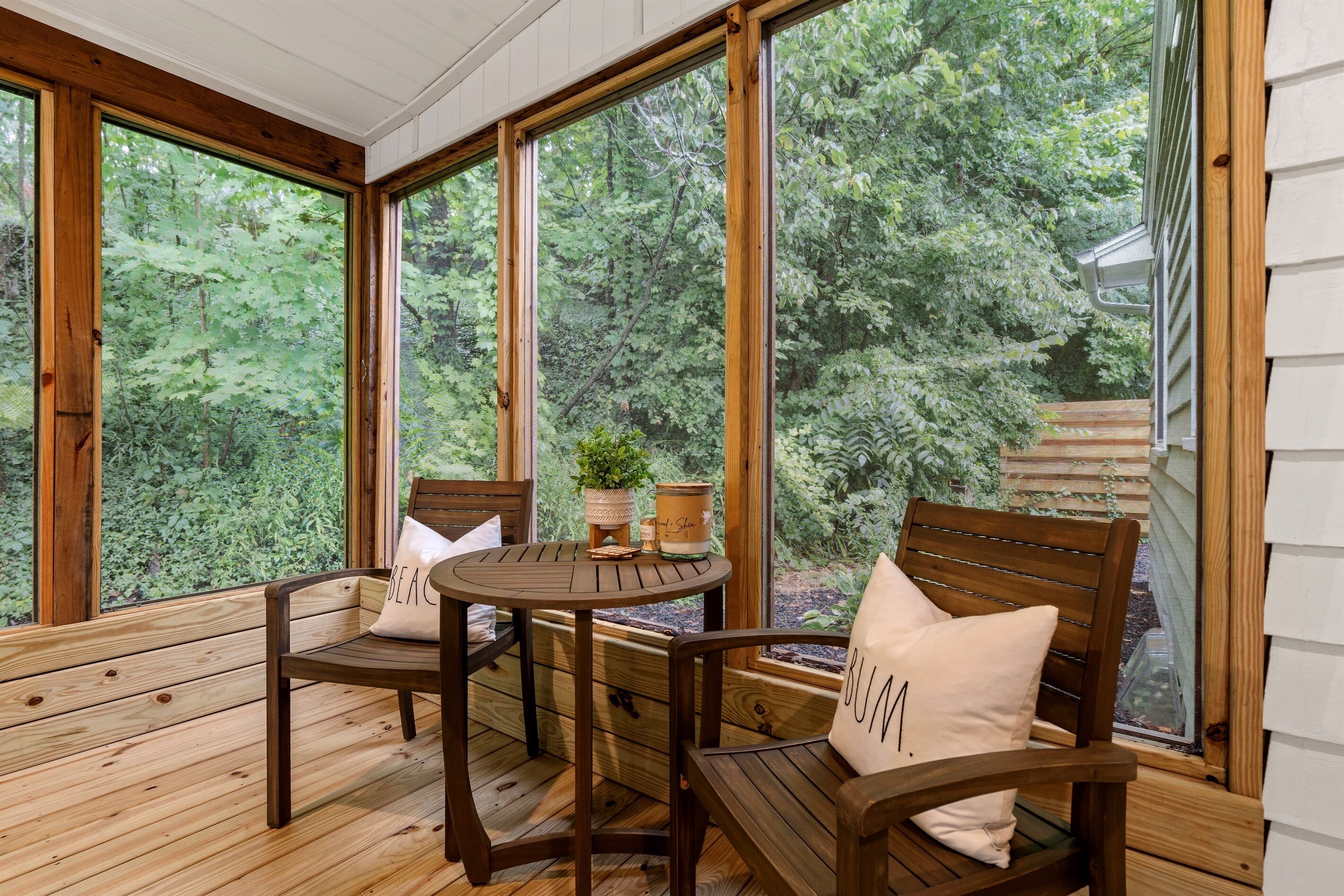 Une terrasse en bois avec une table ronde et deux chaises, de grandes fenêtres donnant sur une forêt, et une petite plante en pot posée sur la table.