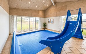 An indoor swimming pool with a blue slide, large windows, and a potted plant.