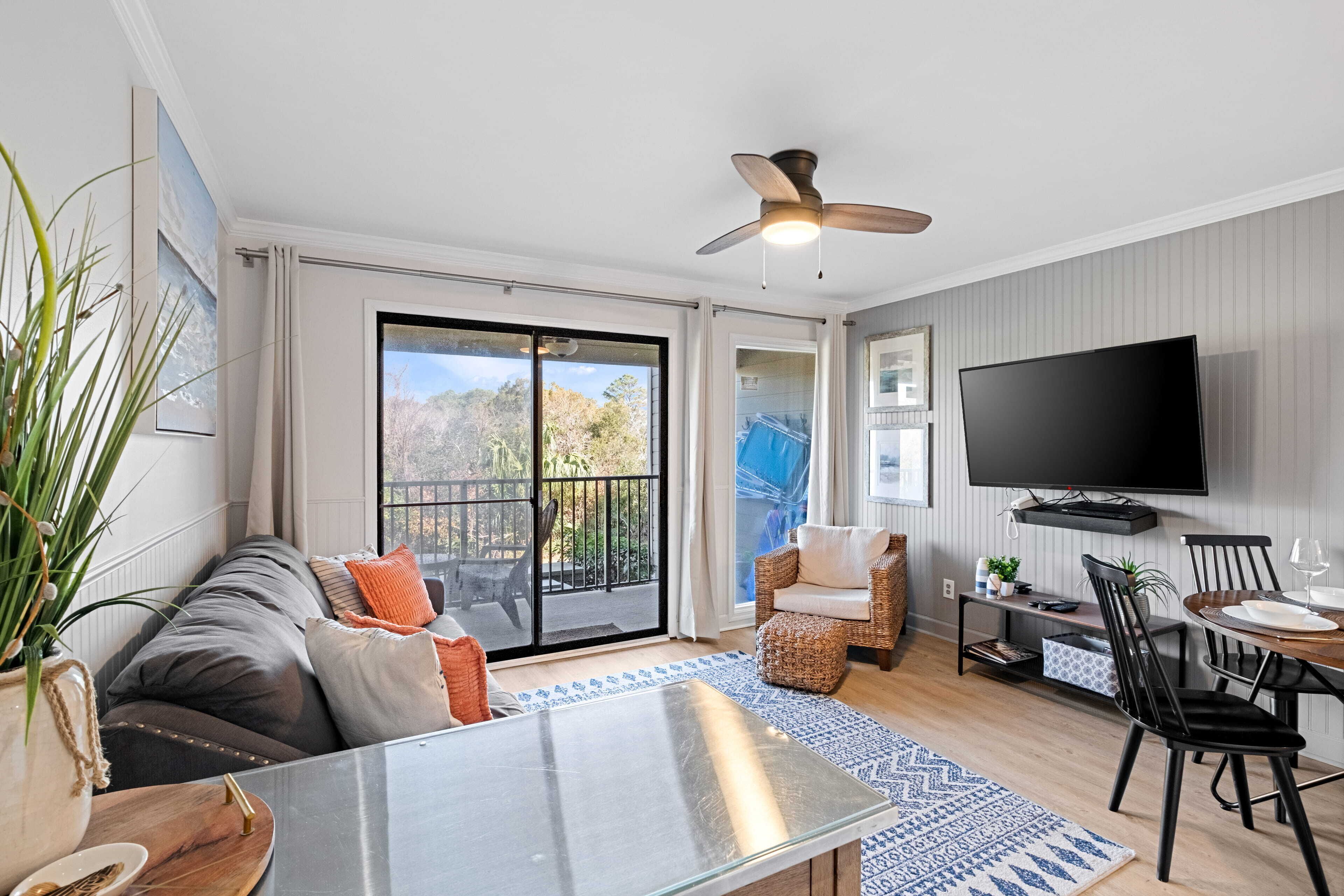 A modern living room with a glass dining table, a sofa with cushions, a ceiling fan, a TV mounted on the wall, and a sliding glass door leading to a balcony.