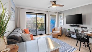Ett modernt vardagsrum med ett glasbord, en soffa med kuddar, en takfläkt, en tv som är monterad på väggen och en skjutdörr i glas som leder ut till en balkong.