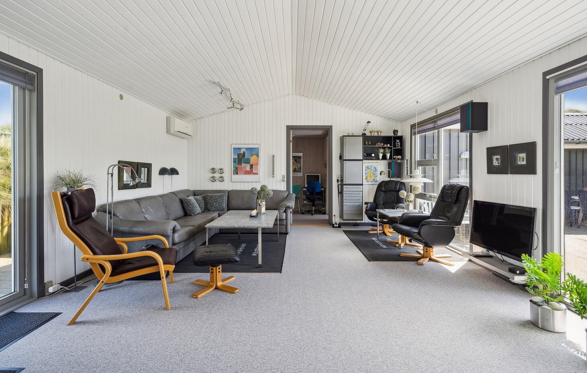 Ein modernes Wohnzimmer mit grauem Sofa, Holzsesseln, einem Couchtisch und einem Fernseher. Es gibt ein Bücherregal mit Dekoration und eine Tür zu einem anderen Raum.