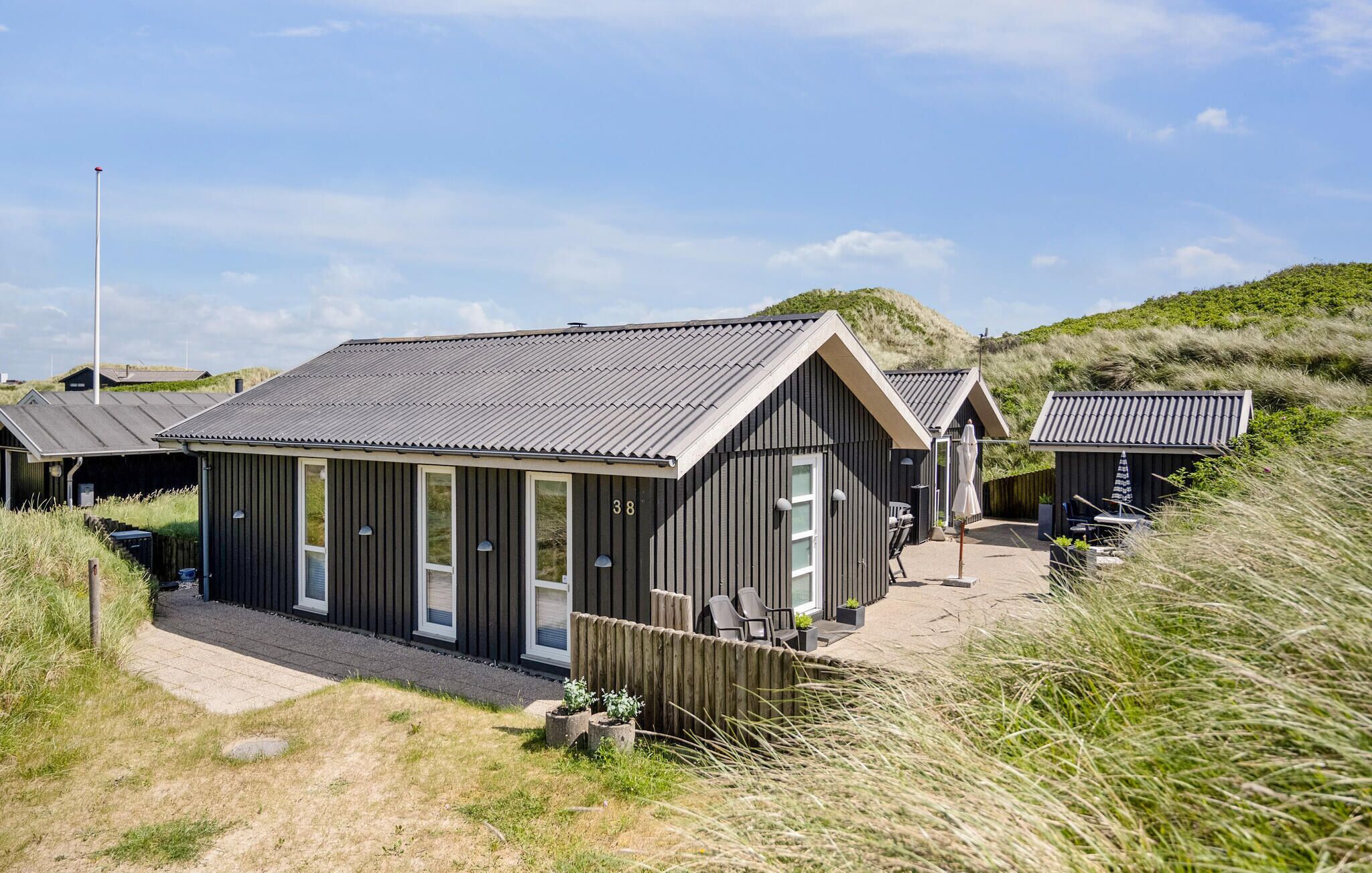 A row of modern, dark-colored cabins with a flat roof, numbered 38, situated in a grassy area with dunes in the background.