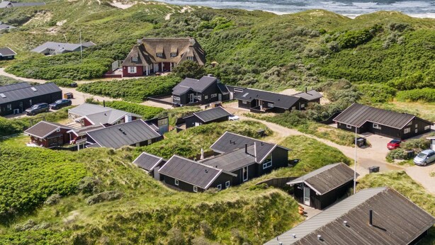An aerial view of a coastal property with multiple buildings, including a prominent house with a thatched roof, surrounded by lush greenery and a sandy beach.