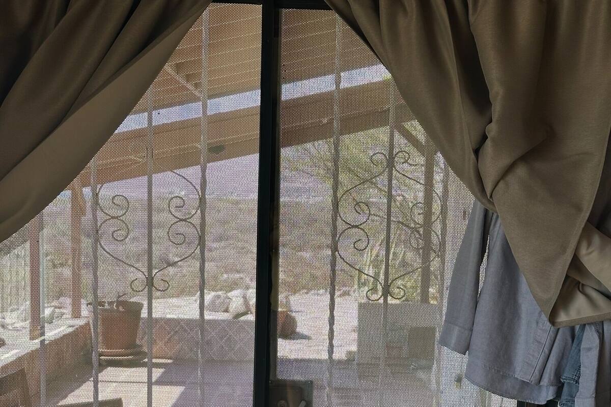 Une porte-fenêtre coulissante avec un moustiquaire métallique, donnant sur un patio extérieur avec une terrasse en bois et des plantes en pot.