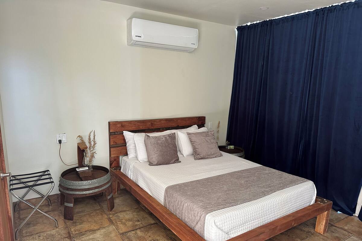 Une chambre à coucher avec un lit en bois, une petite table ronde, un climatiseur fixé au mur et un rideau foncé.