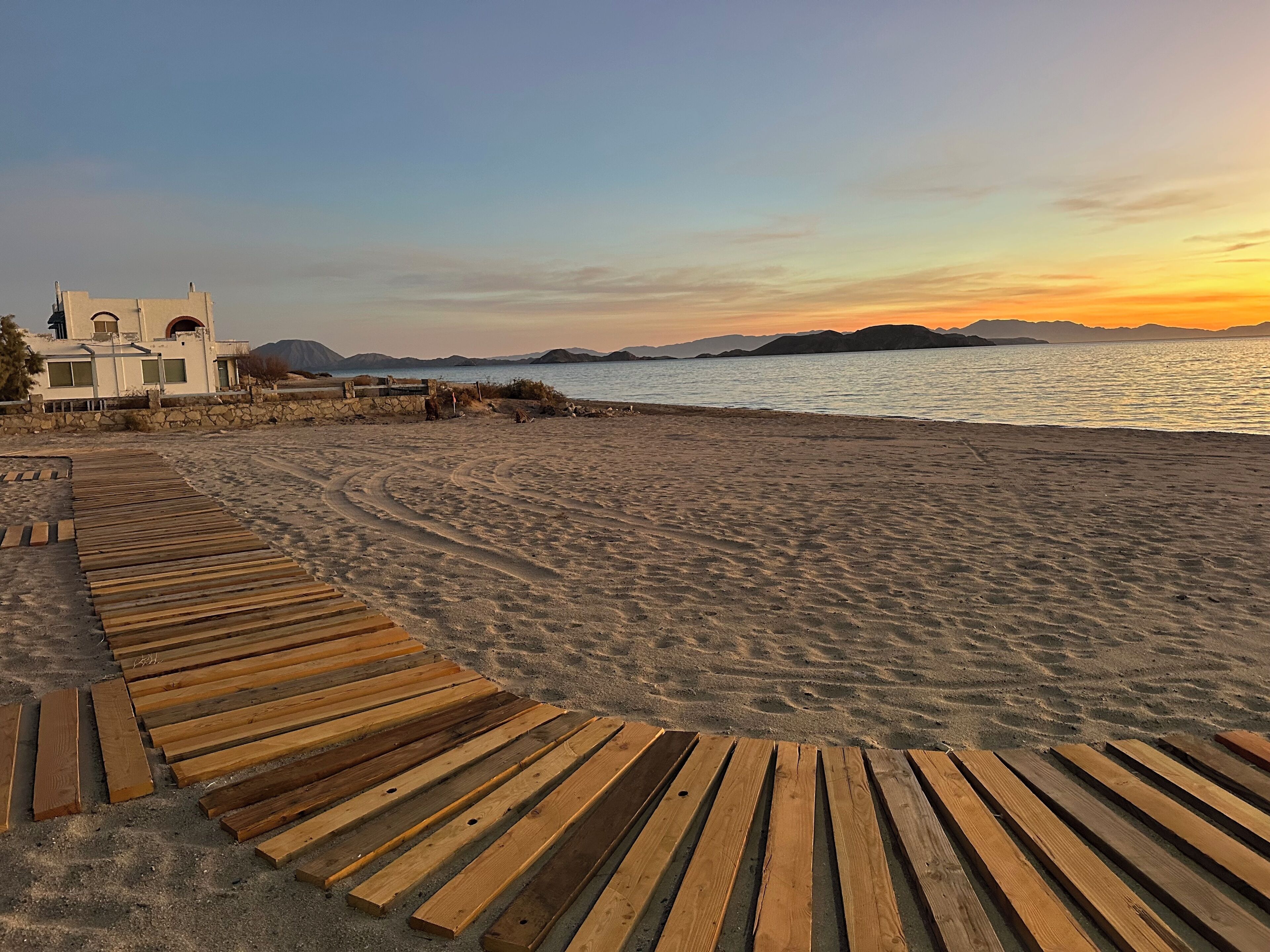 Una terraza de madera que da a una playa, con una casa al fondo.