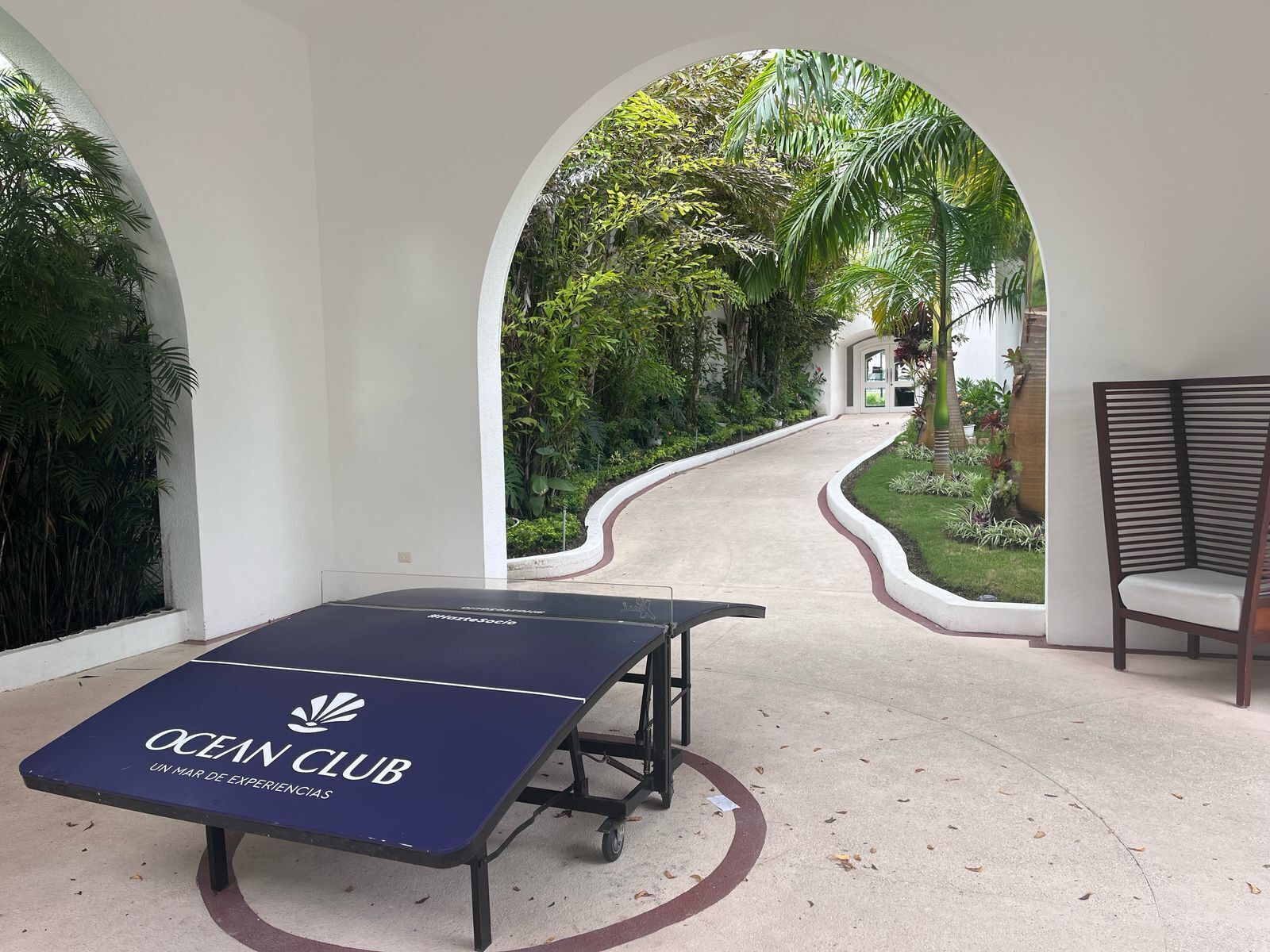 Una mesa de billar con la marca 'OCEAN CLUB' en un área exterior cubierta, con vista a un sendero jardín que lleva a un edificio.