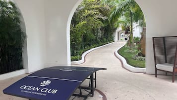 Una mesa de billar con la marca 'OCEAN CLUB' en un área exterior cubierta, con vista a un sendero jardín que lleva a un edificio.