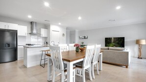 A modern kitchen with white cabinetry, a built-in oven, and a dining area with a wooden table and white chairs. - Seagrass Jervis Bay - Belle Escapes Jervis Bay (Vincentia)
