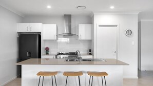 A modern kitchen with white cabinetry, a stainless steel refrigerator, a gas stove, and a central island with bar stools. - Seagrass Jervis Bay - Belle Escapes Jervis Bay (Vincentia)