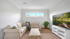 A living room with a beige sofa, a matching ottoman, a large painting of a forest, and a white entertainment unit. - Seagrass Jervis Bay - Belle Escapes Jervis Bay (Vincentia)