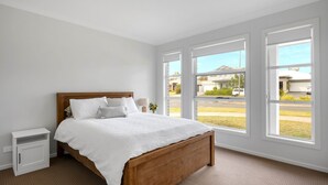 A bedroom with a wooden bed, white bedding, a nightstand, and large windows offering a view of the street. - Seagrass Jervis Bay - Belle Escapes Jervis Bay (Vincentia)