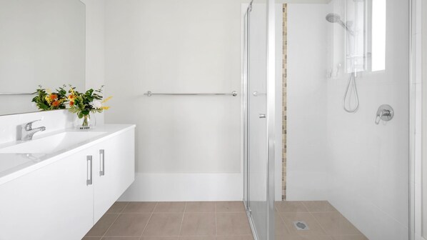 Un baño moderno con cabina de ducha de vidrio, un lavabo blanco y un florero, y piso de baldosas.