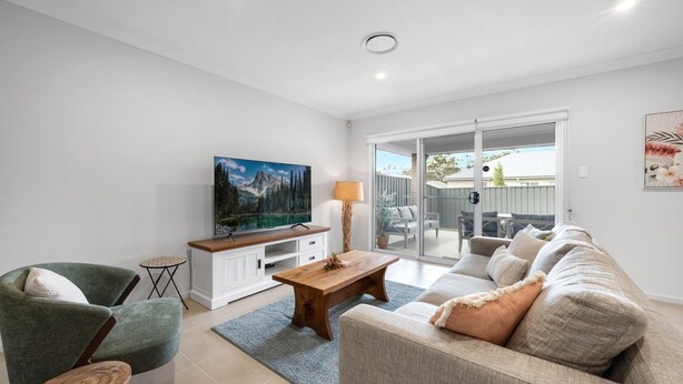 A living room with a flat-screen TV, a wooden coffee table, a sofa, and a chair. There is a sliding glass door leading to an outdoor patio. - Seagrass Jervis Bay - Belle Escapes Jervis Bay (Vincentia)