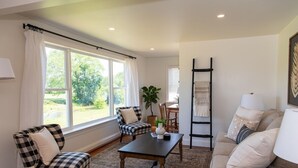 A living room with a checkered armchair, a sofa, a coffee table, and a view of the outdoors through large windows. - Amish Cottage, Hot Tub, on Mill Creek (Lancaster)