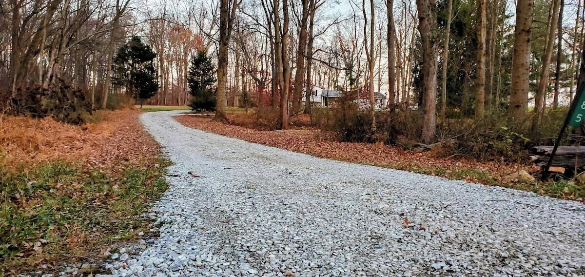 Een grindpad in een bebost gebied met bomen en een huis op de achtergrond.