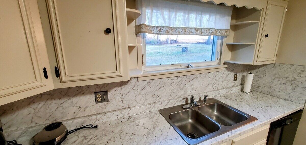 Una cocina con un lavamanos doble, armarios blancos, un ventanal con adornos y una encimera de mármol.