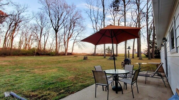 A patio with a table and chairs, an umbrella, and a view of a grassy yard and trees.
