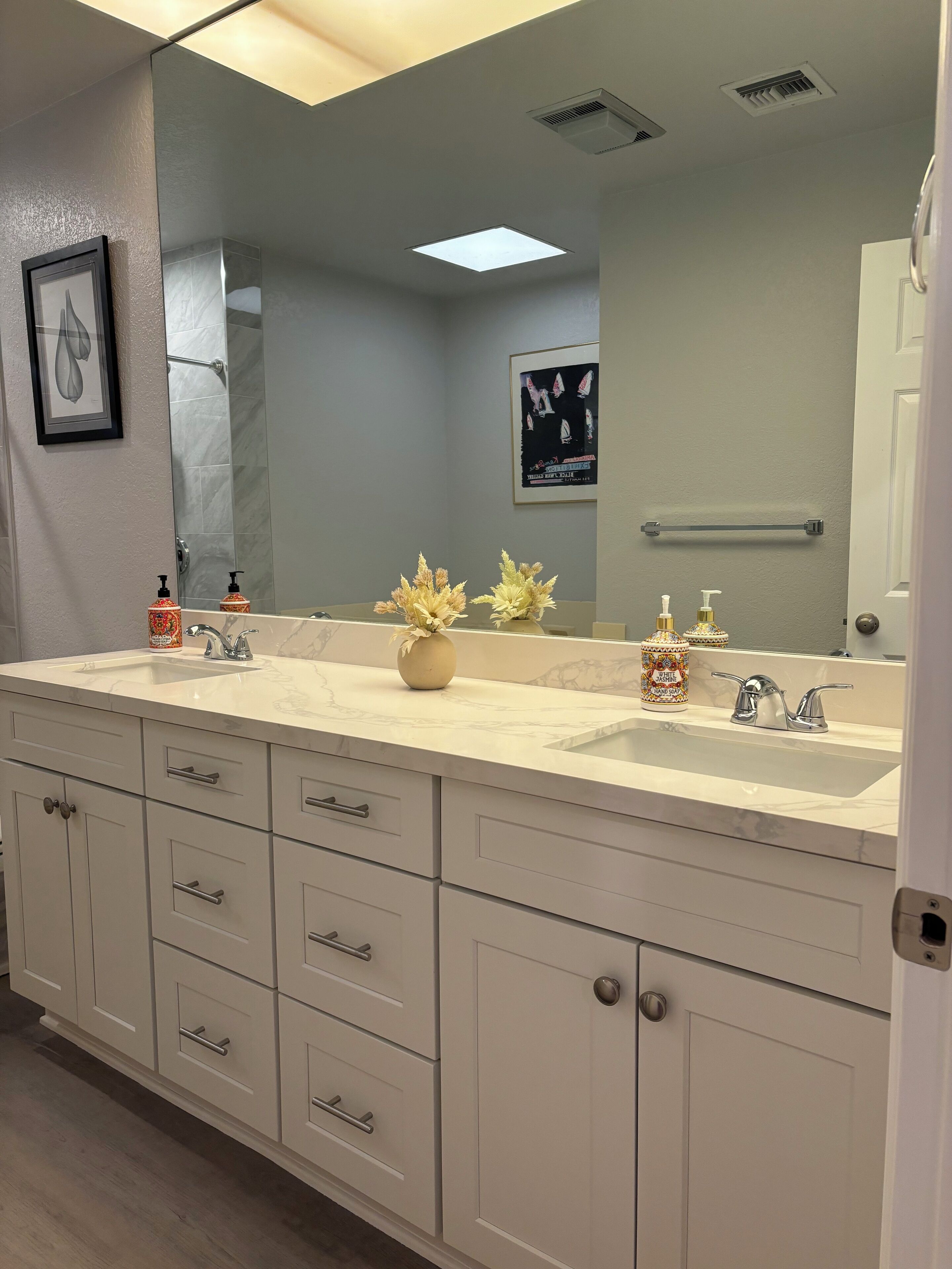 A bathroom with a large mirror, a sink, and a vanity with drawers. There are two soap dispensers on the sink, and a vase with flowers on the vanity.