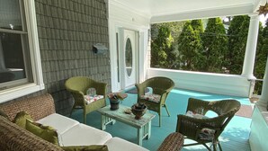 A covered porch with wicker furniture, a small table with a plant, and a view of trees. - Charming 1-bedroom apartment - 38 Mins to NYC / Parking on site (New Rochelle)