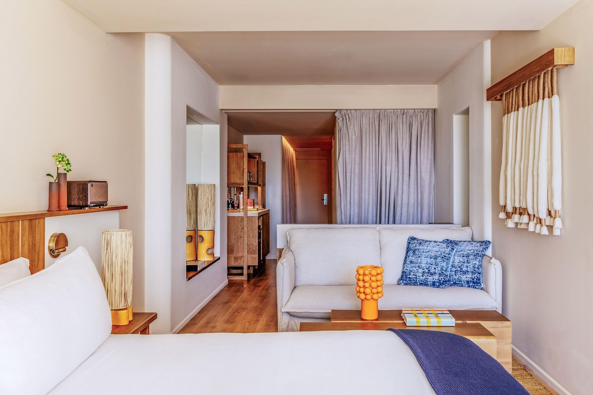 A hotel room with a bed, a sofa, a coffee table, a lamp, a mirror, and a wardrobe.