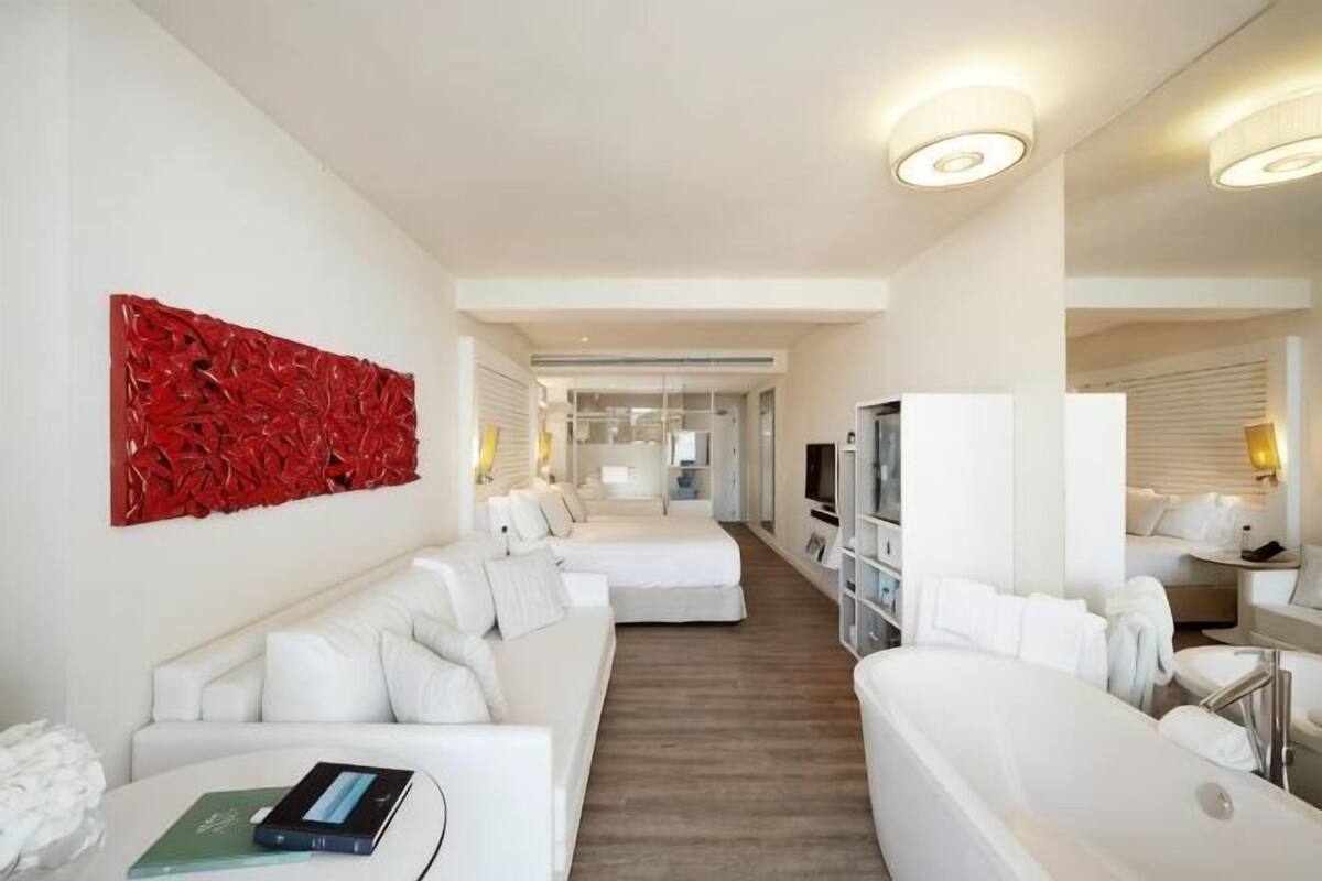 Une chambre d’hôtel moderne avec un canapé blanc, une grande œuvre d’art abstraite rouge, une baignoire et un couloir menant à une autre pièce.