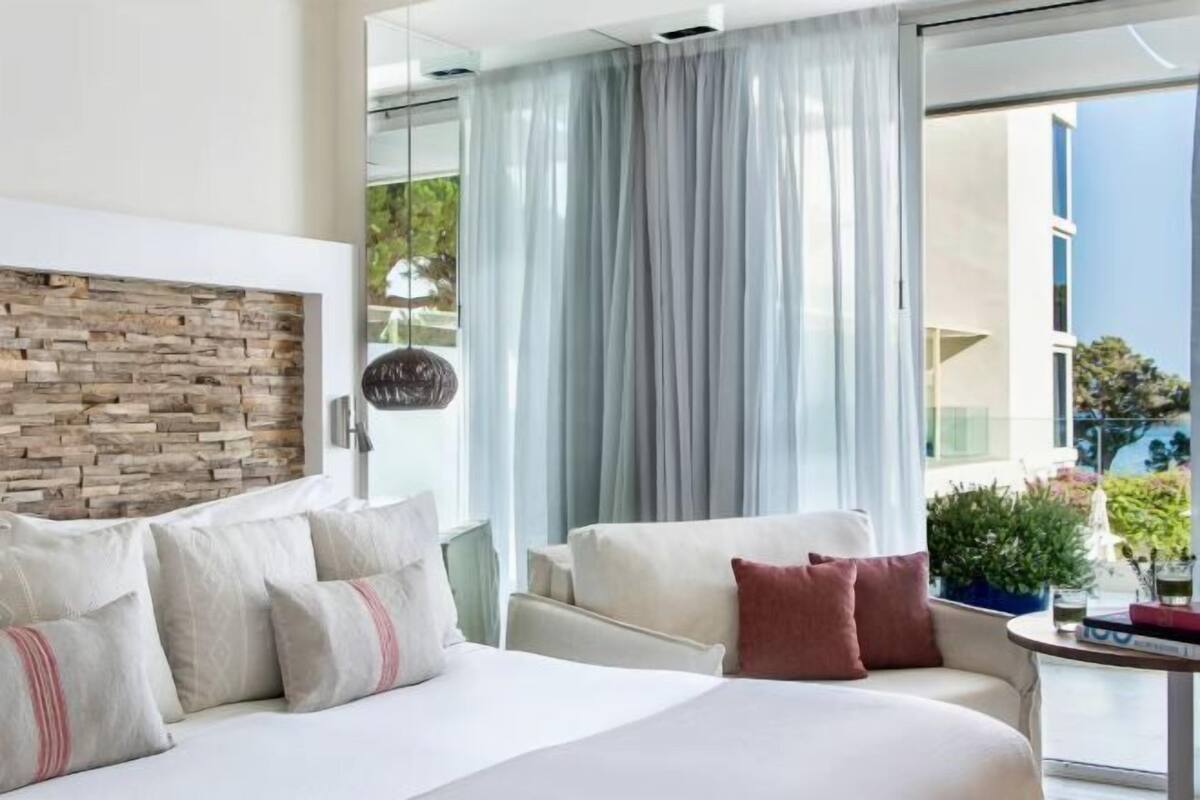 Une chambre d’hôtel moderne avec un mur d’accent en pierre, un lit agrémenté de plusieurs oreillers, un canapé et une vue sur la verdure environnante.