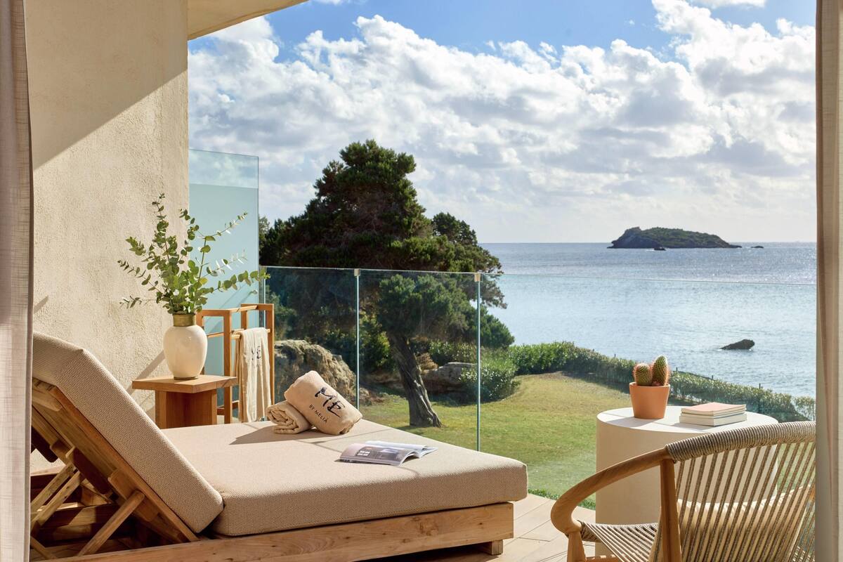 Un balcon doté d’un espace salon avec des coussins, d’une petite table sur laquelle se trouve un livre et un pot de fleurs, et d’une vue sur la mer et une île lointaine.