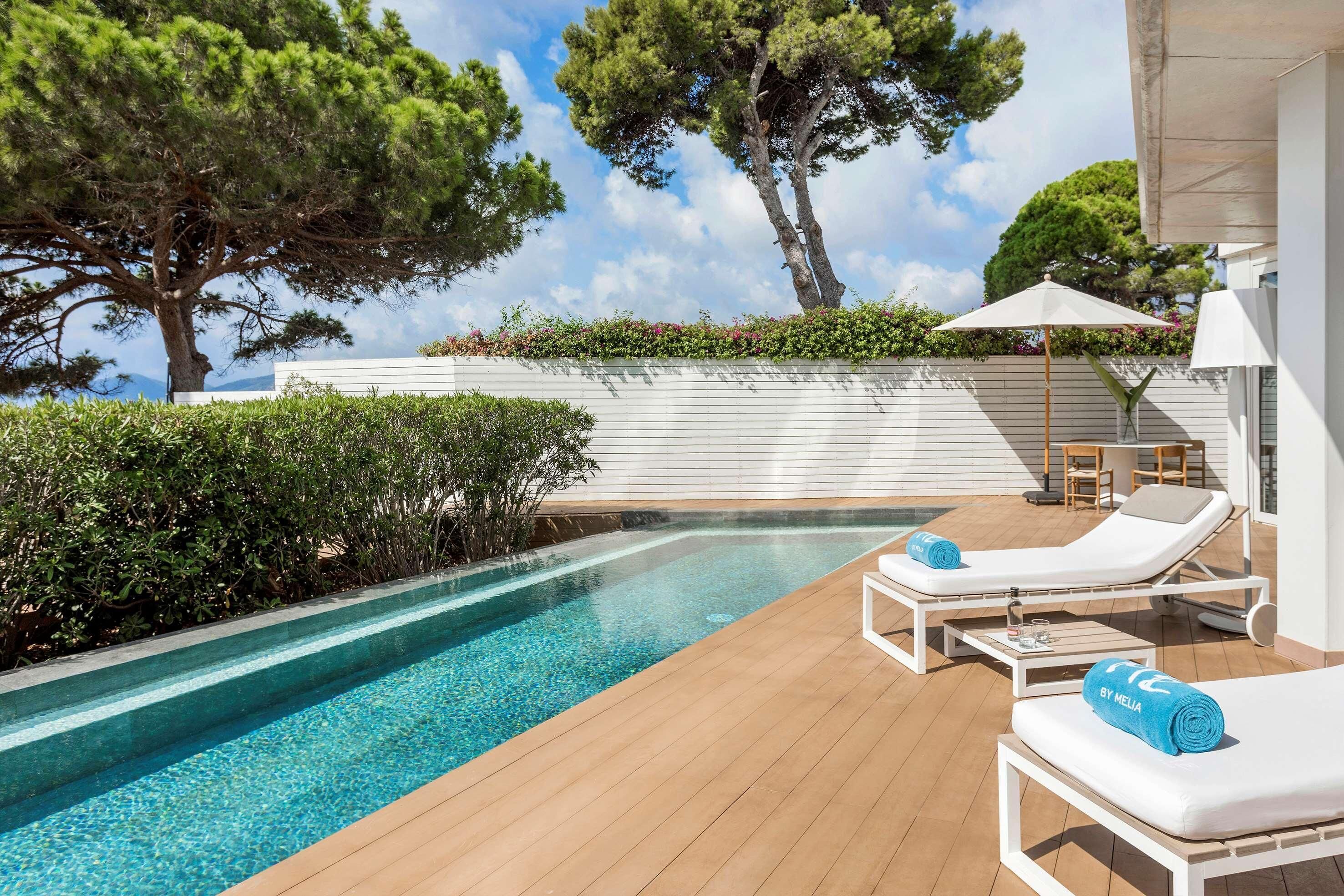 A pool area with lounge chairs, a wooden deck, and a white wall with greenery.