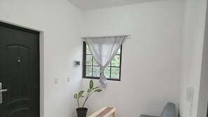 A room with a black door, a window with a curtain, a potted plant, a wooden bench, and a grey sofa. - Apartamento #3 (Macario Gómez)
