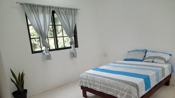 A bedroom with a bed, a window with curtains, a potted plant, and a wall socket. - Apartamento #3 (Macario Gómez)