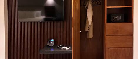 A modern room with a flat-screen TV mounted on a wooden wall, a small desk with a phone and remote controls, and a wooden wardrobe with a hanging garment.