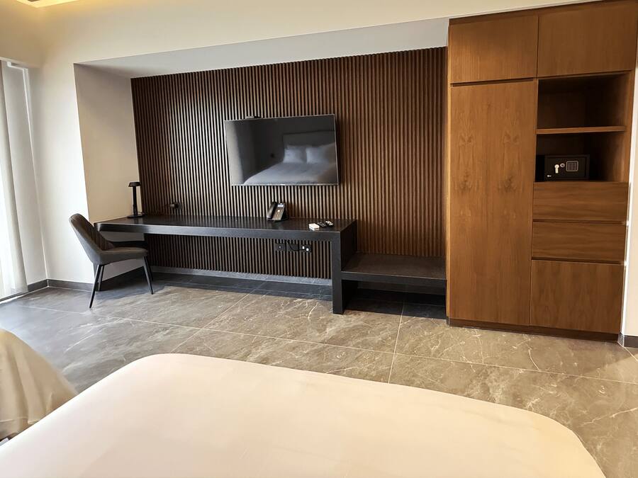 Una habitación de hotel moderna con una cama grande, un escritorio con una silla, un televisor de pantalla plana montado en una pared de paneles de madera y un armario de madera.