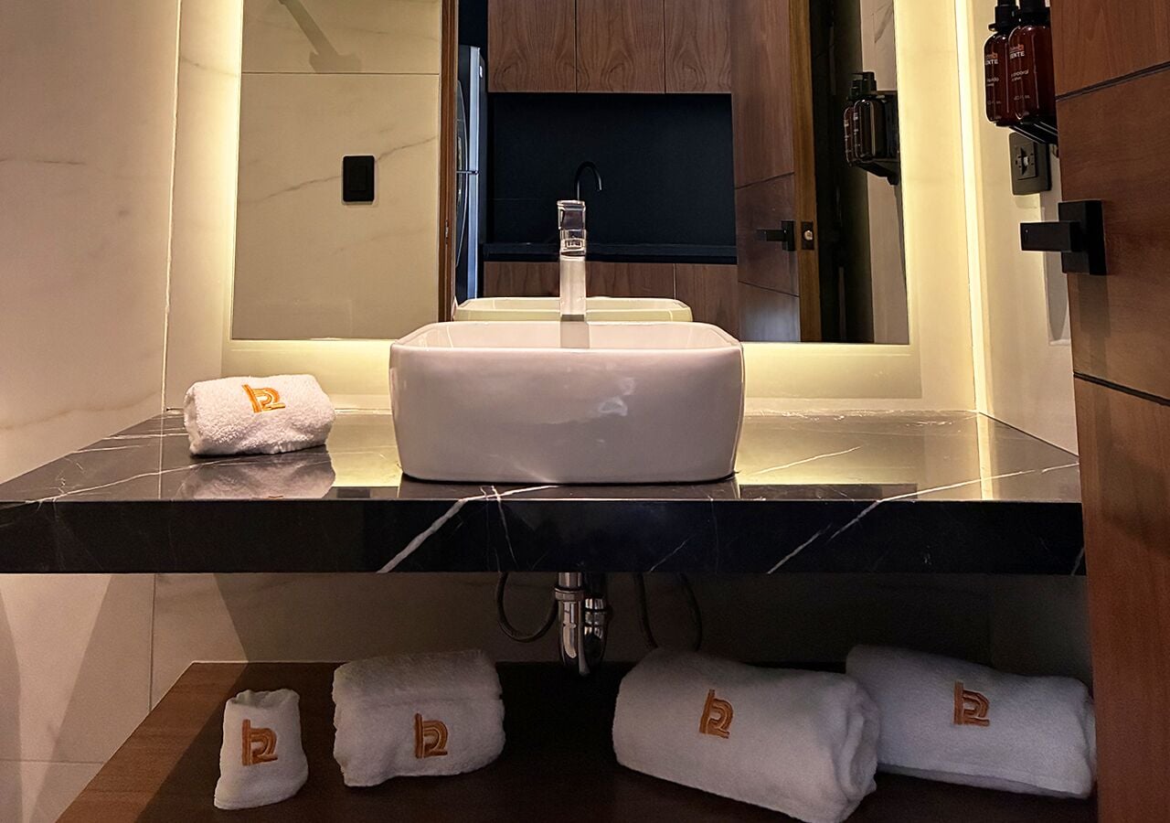 Une salle de bain moderne avec un lavabo central, un miroir au-dessus, et des serviettes ornées d’un logo, disposées sur le comptoir et sur le sol.