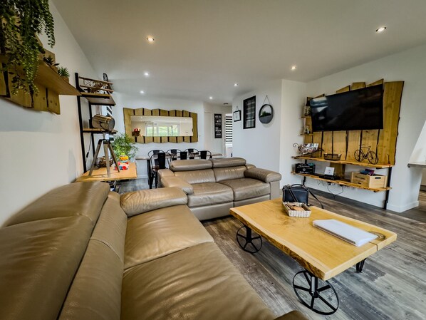 Een moderne woonkamer met een hoekbank, een eethoek en een televisie bevestigd aan een houten wand.