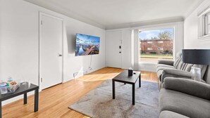Een moderne woonkamer met een flatscreen televisie aan de muur, een houten vloer, een grijze bank, een klein zwart tafeltje en een groot raam met uitzicht op een bakstenen gebouw.