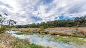 A river flowing through a natural landscape with trees and vegetation on the banks. - 6) Texas Pride  at Vineyard Trail Cottages (Fredericksburg)