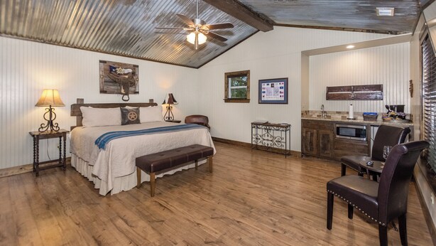 A bedroom with a bed, bedside tables, a bench, a ceiling fan, and a kitchenette with a microwave. - 6) Texas Pride  at Vineyard Trail Cottages (Fredericksburg)