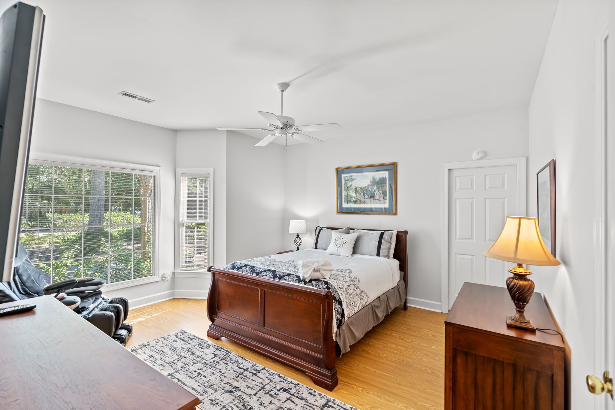 A bedroom with a wooden bed, a ceiling fan, a bedside lamp, a framed picture, and a window with blinds.