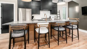 Uma cozinha moderna com armários escuros, uma geladeira de aço inoxidável, uma ilha central com banquetas, iluminação pendente e uma janela grande.