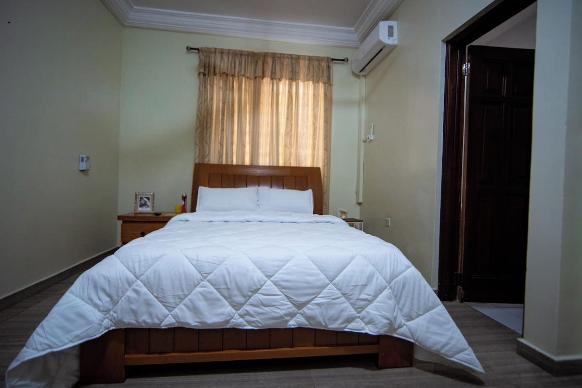 A neatly made bed with a wooden frame and white bedding, a bedside table with a lamp, and a window with curtains.