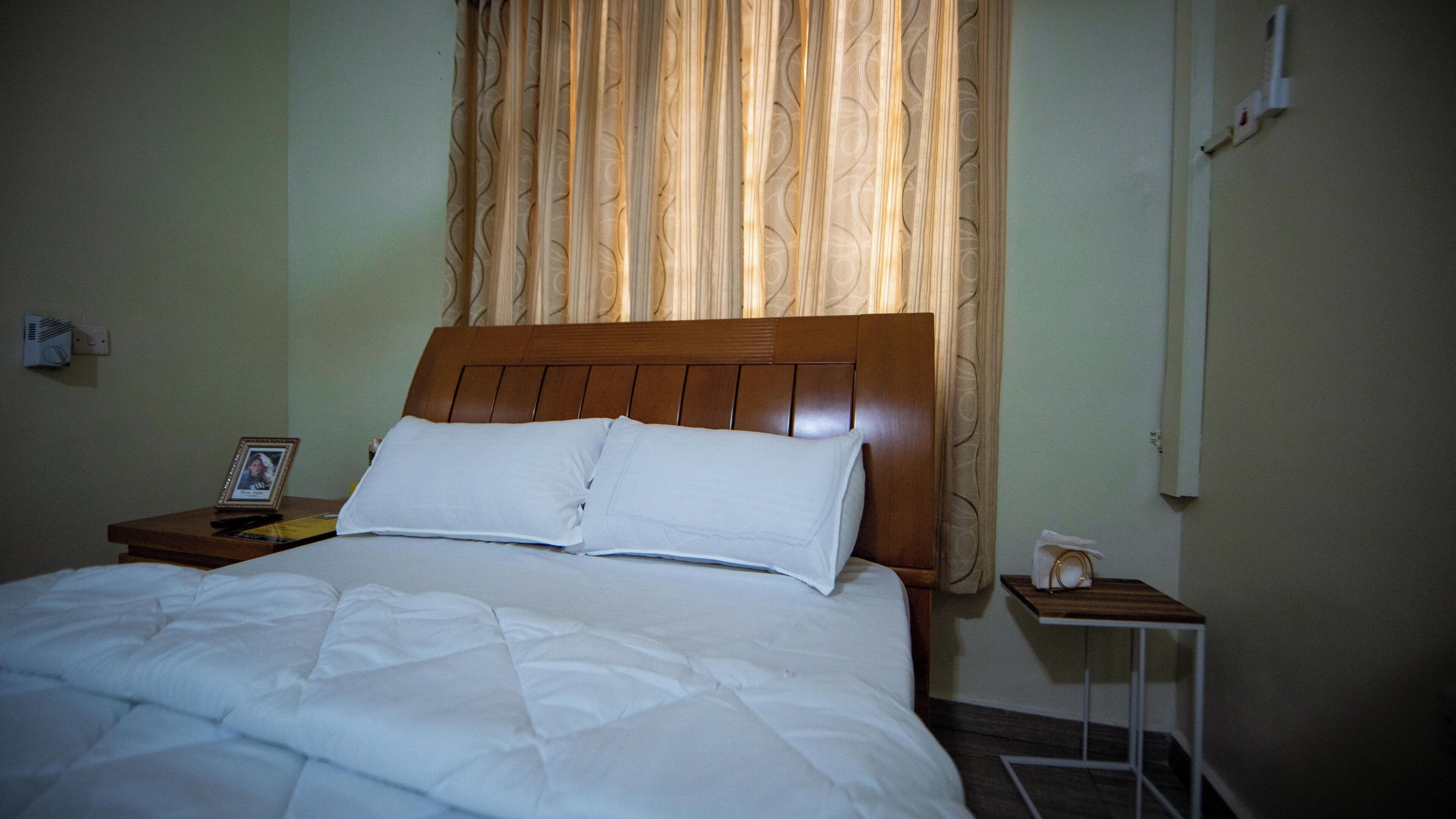 Una cama bien hecha con ropa blanca, un cabecero de madera y una mesita de noche con una foto enmarco y un control remoto.