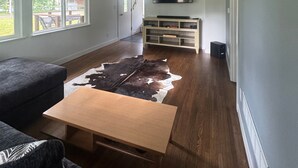 En rummelig stue med et stort vindue, et træsofabord, en lædermatte og et tv ophængt på væggen.