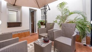 Un espacio al aire libre acogedor con mobiliario de mimbre, una mesa con tetera y un gran paraguas que ofrece sombra.