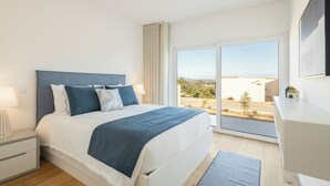 Ein modernes Schlafzimmer mit einem großen Bett, einem Nachttisch, einer Lampe und Blick ins Freie durch ein großes Fenster.