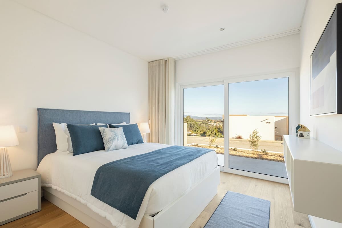 Quarto moderno com uma cama grande, uma mesinha de cabeceira, uma luminária e vista para o exterior através de uma janela ampla.