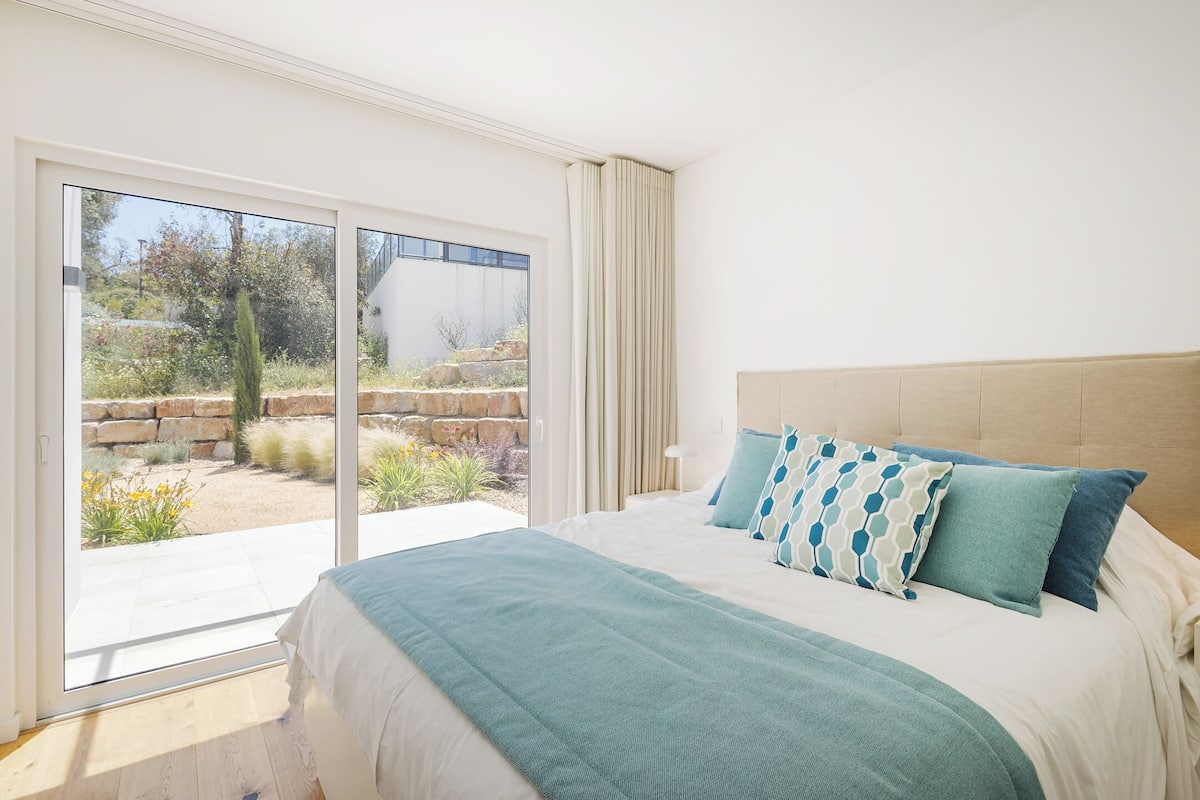 Quarto com cama grande, vista para um jardim através de portas de correr de vidro e cabeceira bege.
