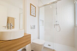 Un baño moderno con lavamanos blanco, tocador de madera, inodoro y zona de ducha con mampara de vidrio.