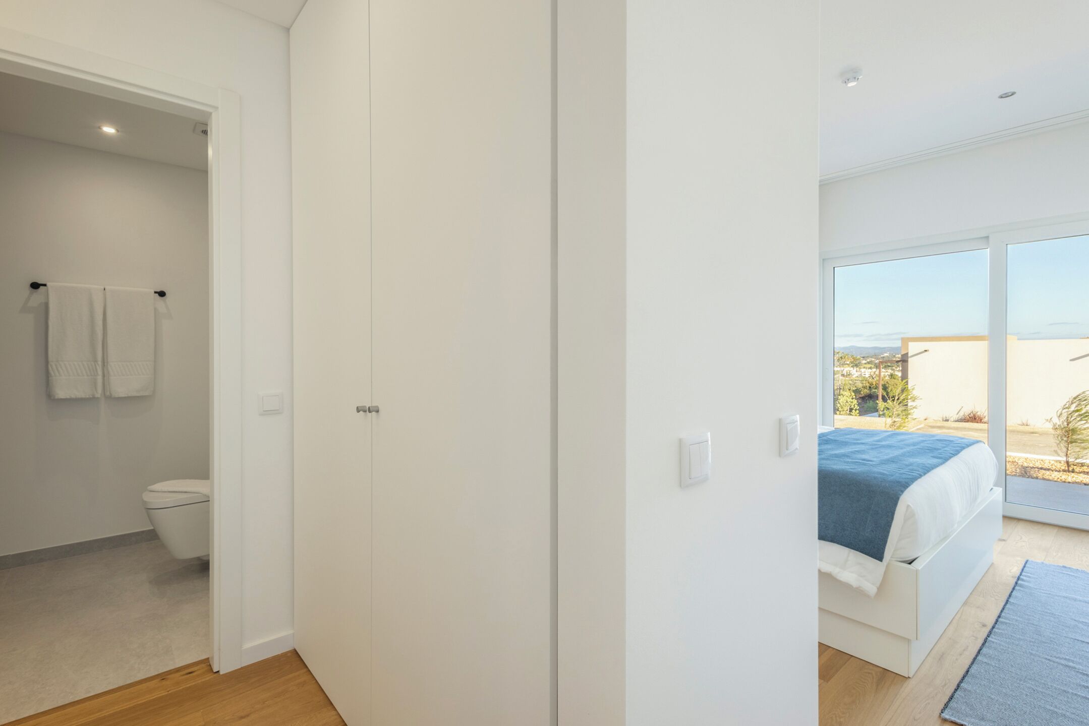 A bedroom with a bed, a view through a large window, and a bathroom with a toilet and towel rack.